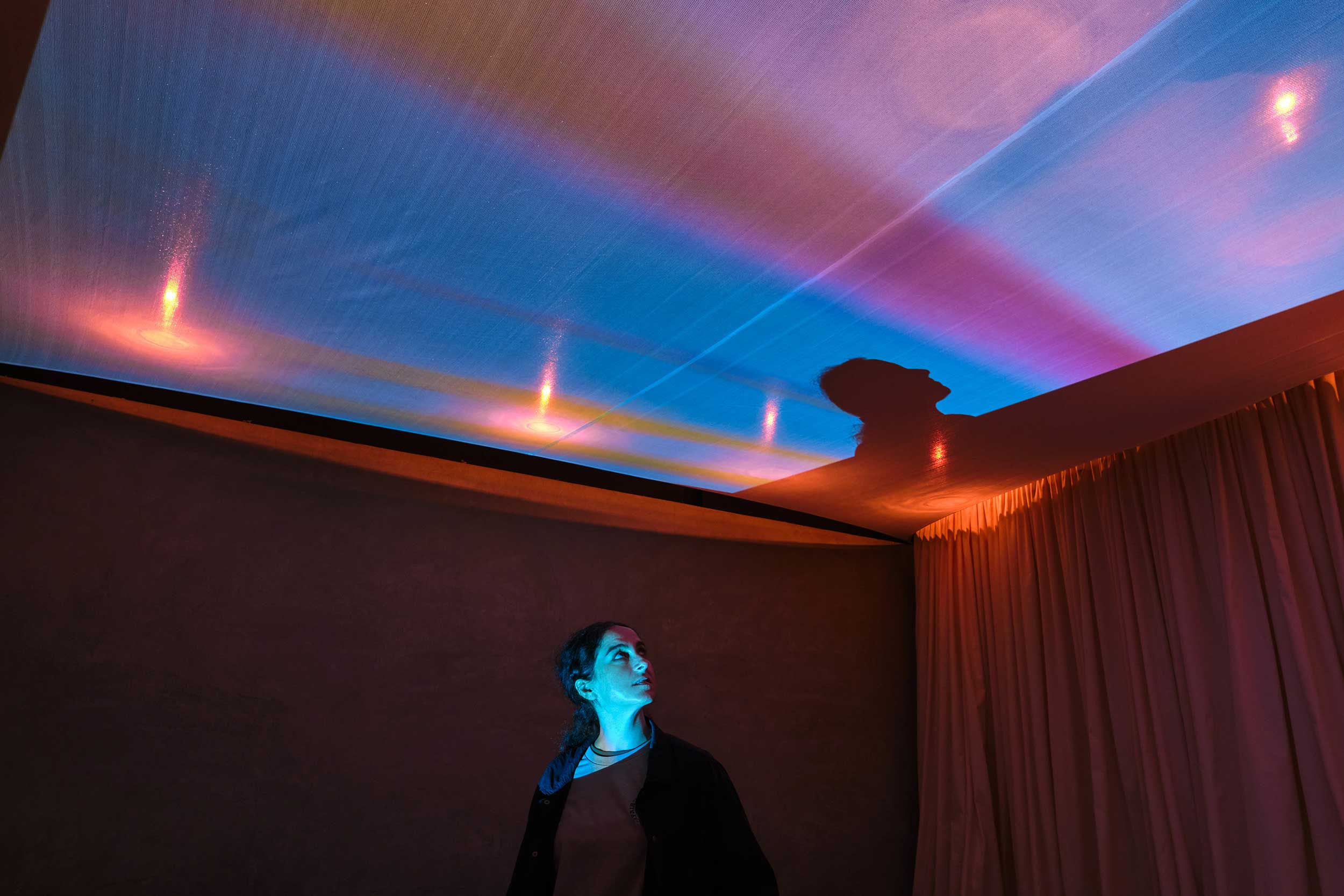 A woman stands in a dark space with her face illuminated by video projection on the ceiling