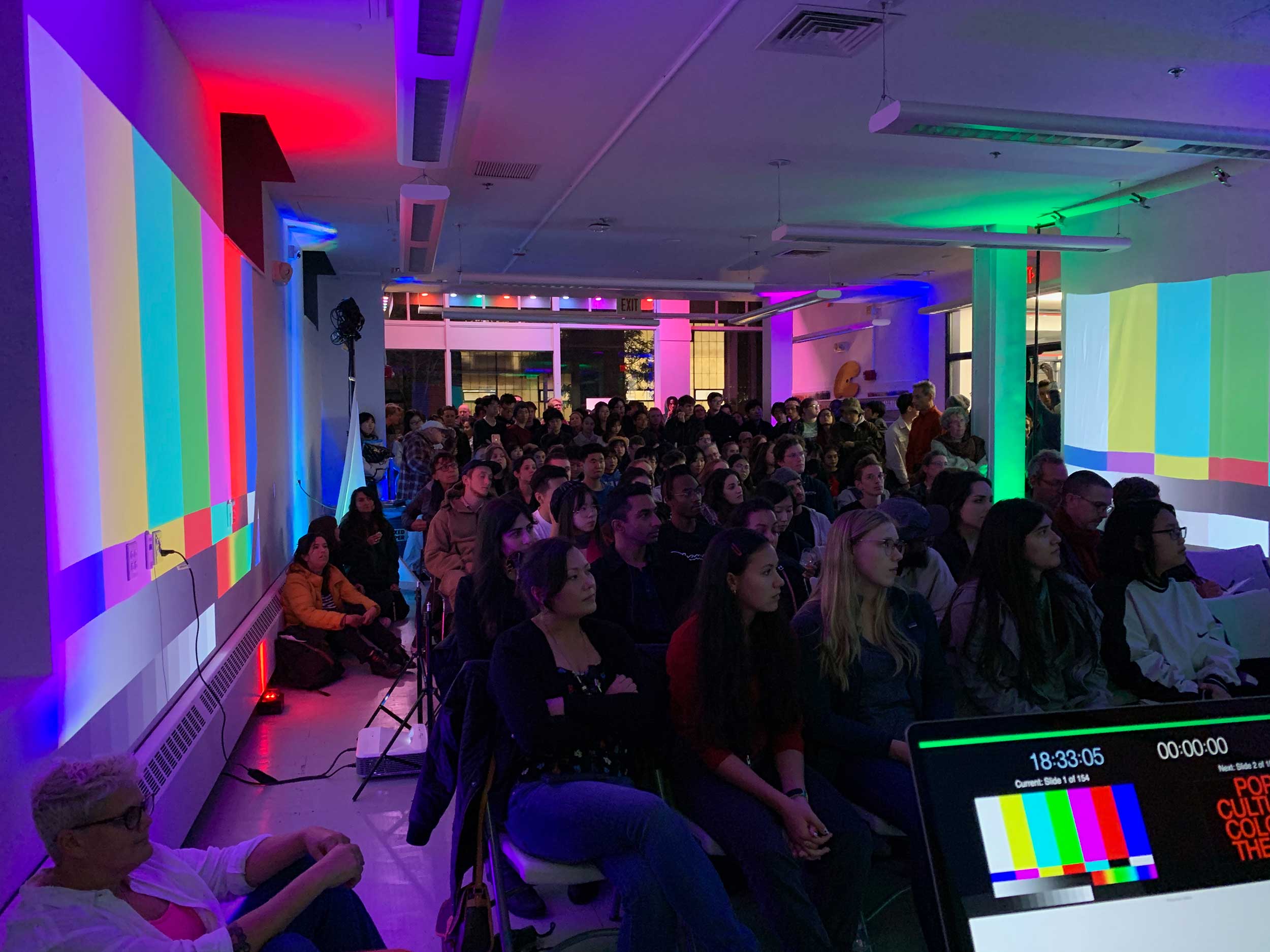A crowded lecture with multi-colored test pattern projections on both side walls and colored lights in the background