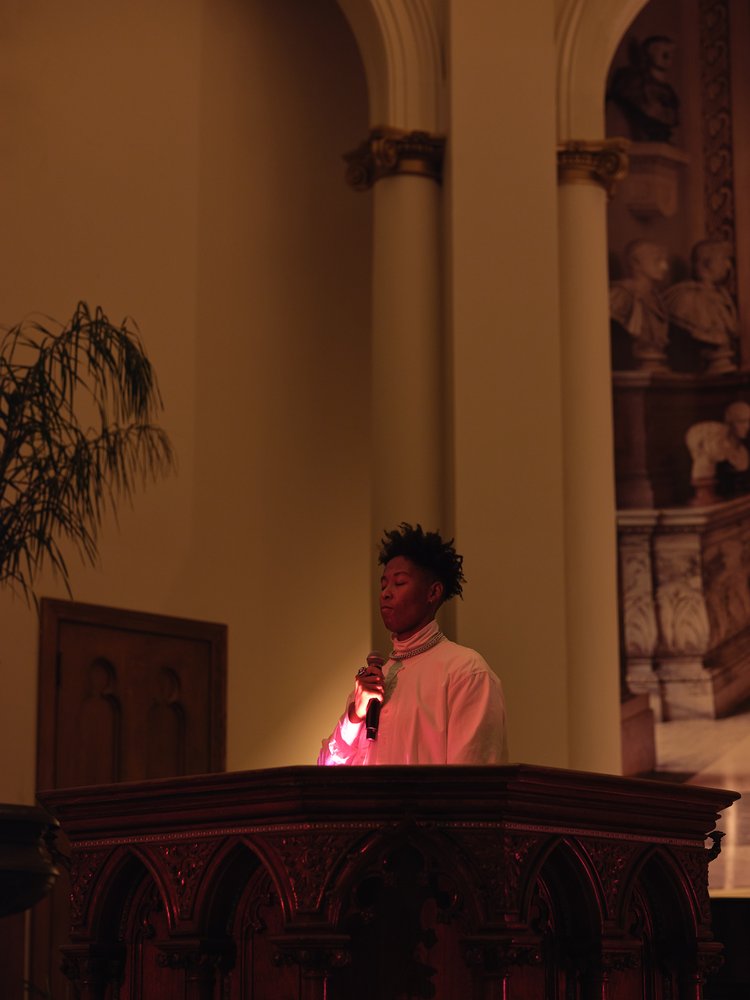 Artist Leila Weefur holding a microphone and standing at a pulpit lit by pink light