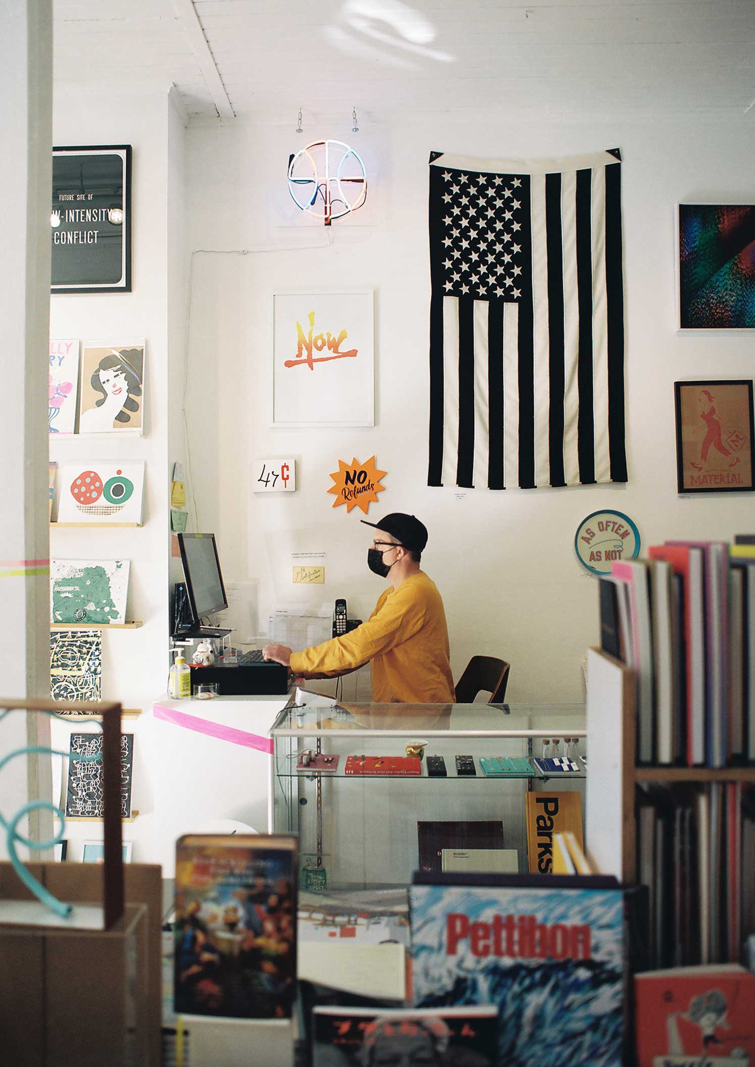 Man in yellow sweater standing at retail desk surrounded by black-and-white US flag, graphic posters, and books
