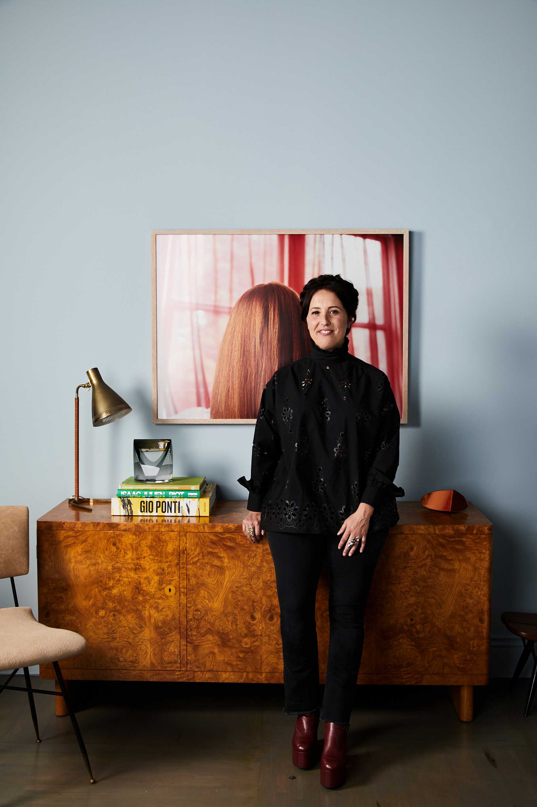 A woman dressed in black standing in front of a wood credenza, red photographic artwork, and pale blue wall