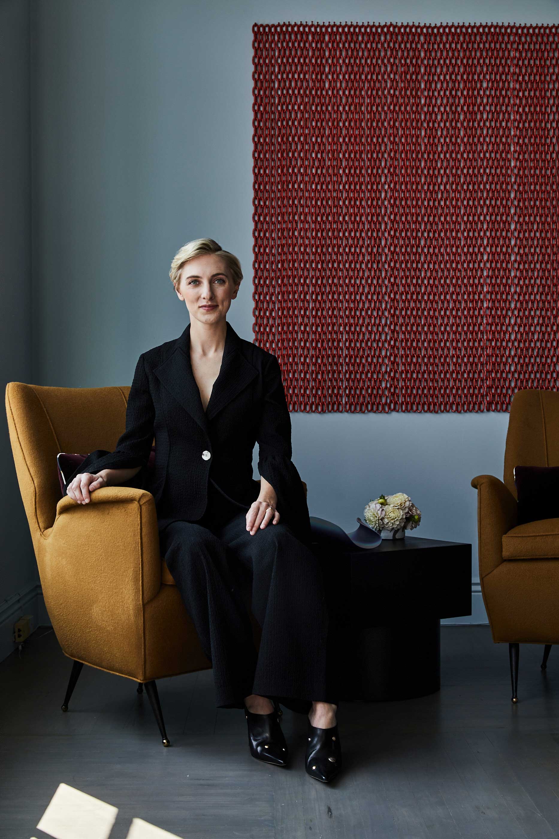 A woman dressed in black sitting on a dark yellow lounge chair in front of a red artwork hanging on a pale blue wall
