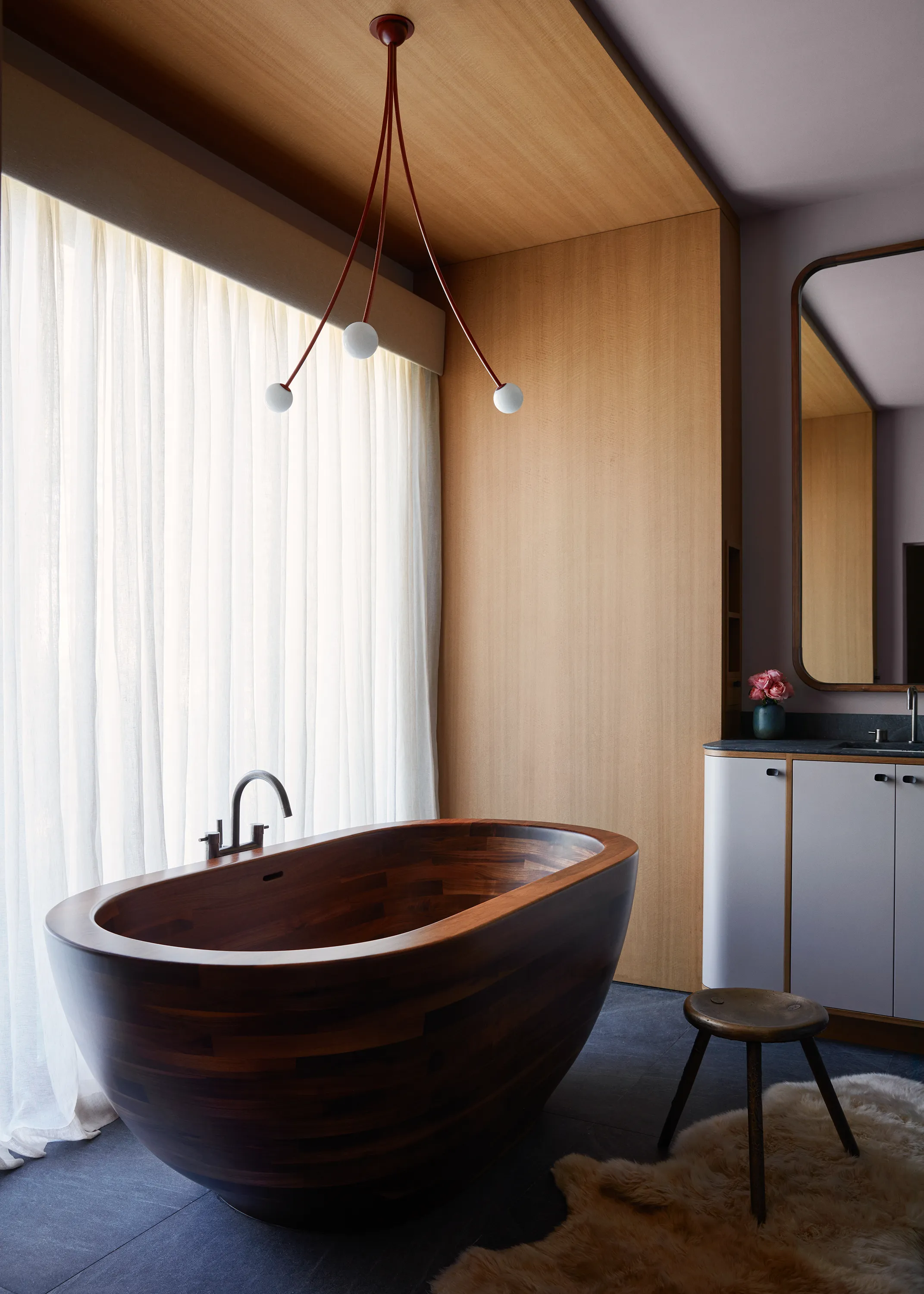 A walnut Karolina bathtub by Aquatica basks beneath a Michael Anastassiades Fontana Amorosa Bouquet Ceiling Pendant attended by a bronze stool by The Future Perfect.