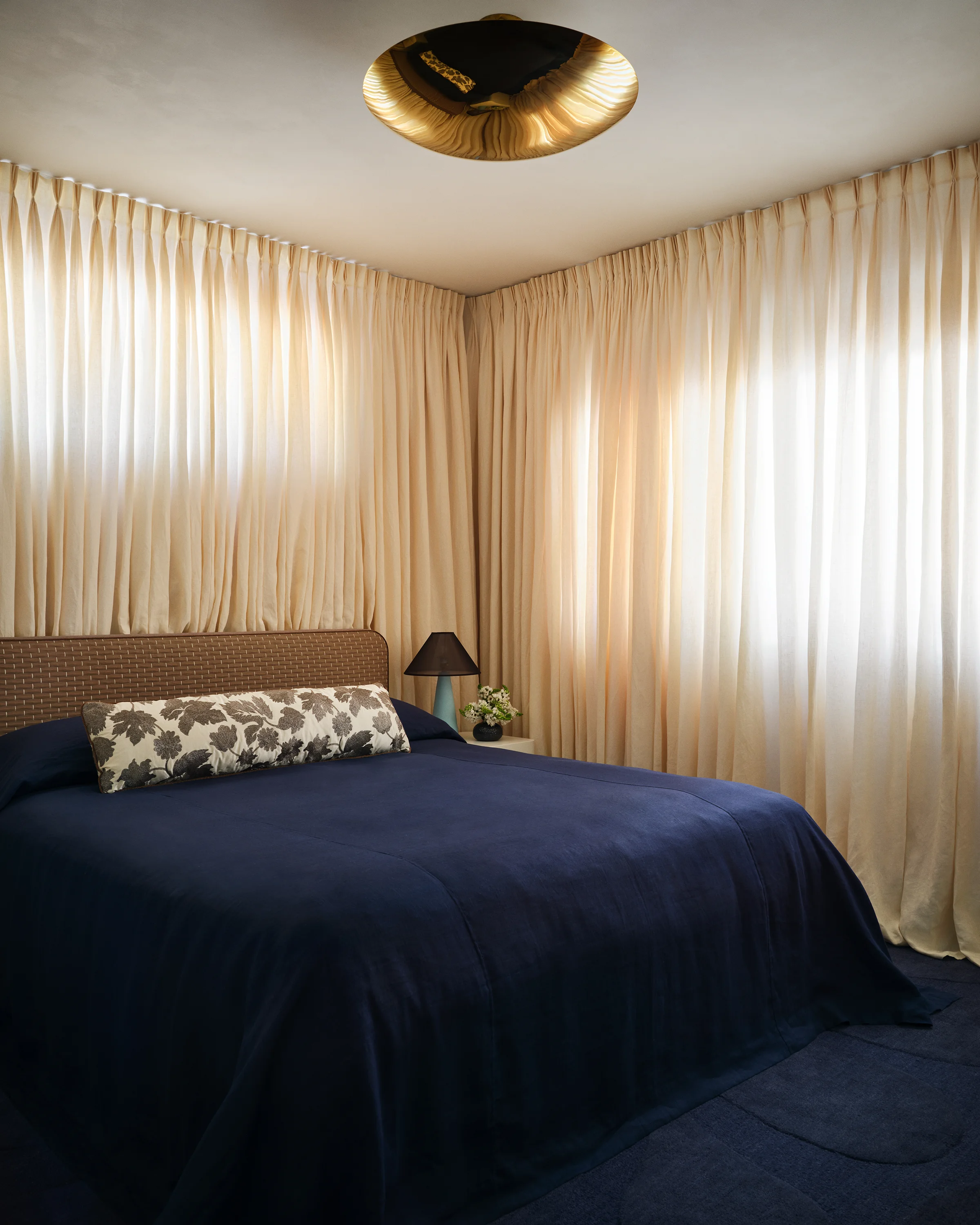 A dramatic brass ceiling fixture crowns a woven headboard with navy bedding and botanical pillow beneath floor-to-ceiling sheer curtains in contemporary Sonoma guest bedroom.