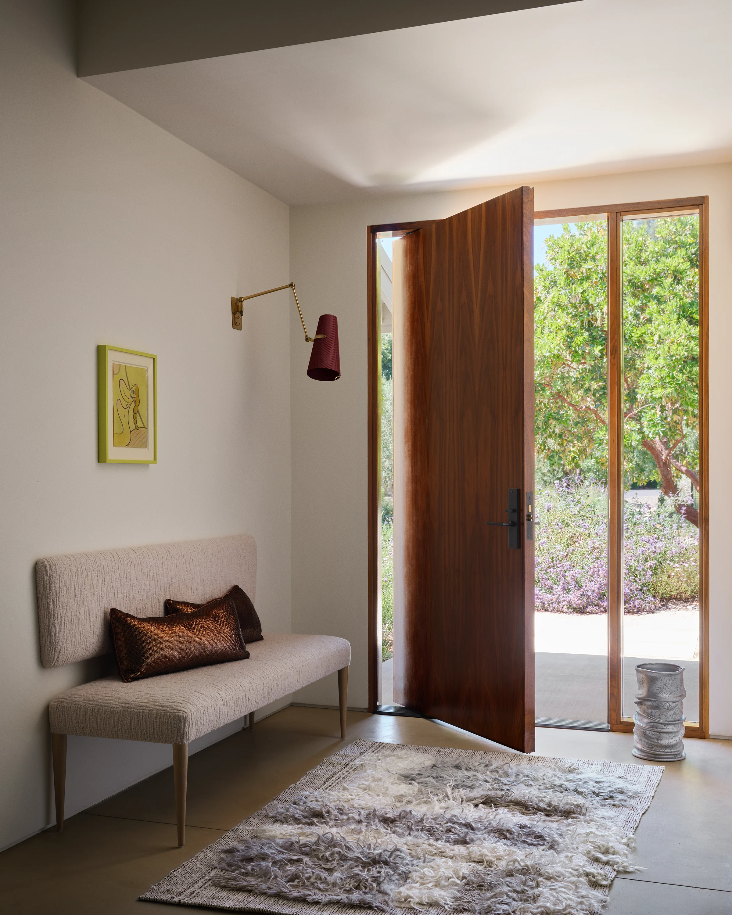 A sculptural walnut door opens to reveal the landscape design beyond as vintage brass sconce and line drawing welcome guests at a neutral bench with textured pillows in contemporary Sonoma entry.