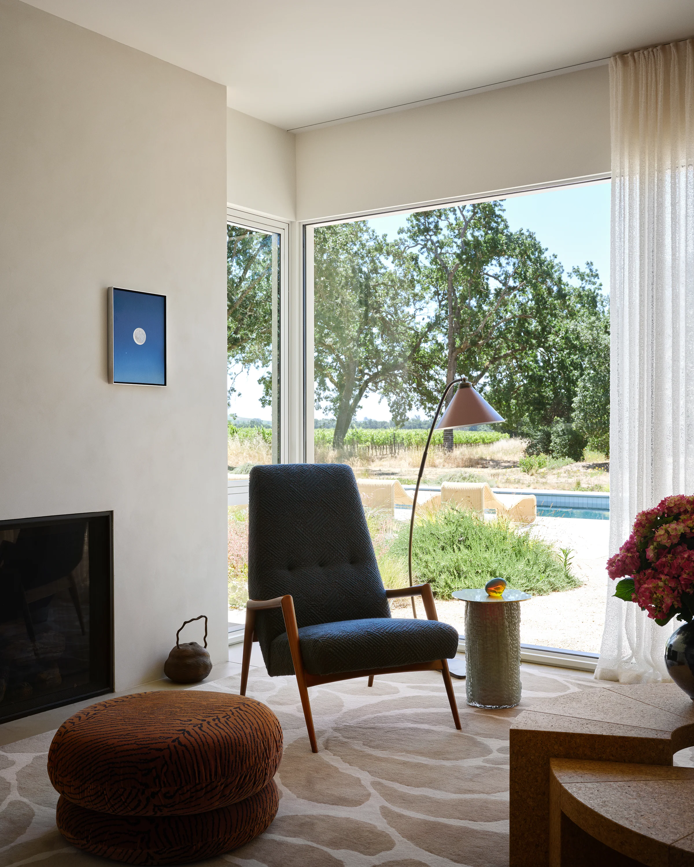 Vintage high-back chair and woven ottoman frame vineyard views beside minimalist fireplace with contemporary moon painting in serene Sonoma living room.