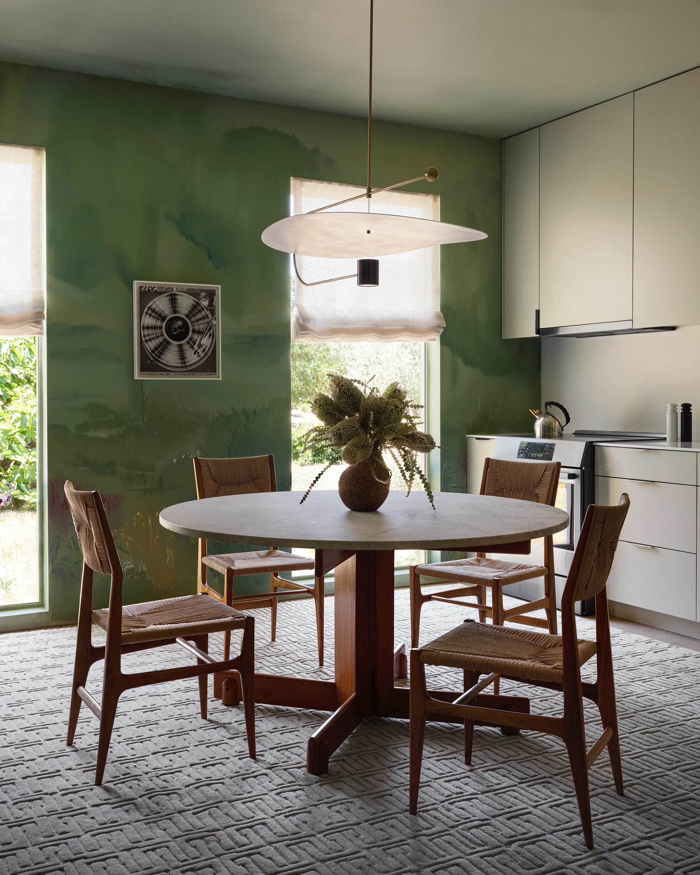 Vintage woven dining chairs encircle a round vintage table beneath a sculptural pendant light against sage green mural walls in contemporary Sonoma dining room.