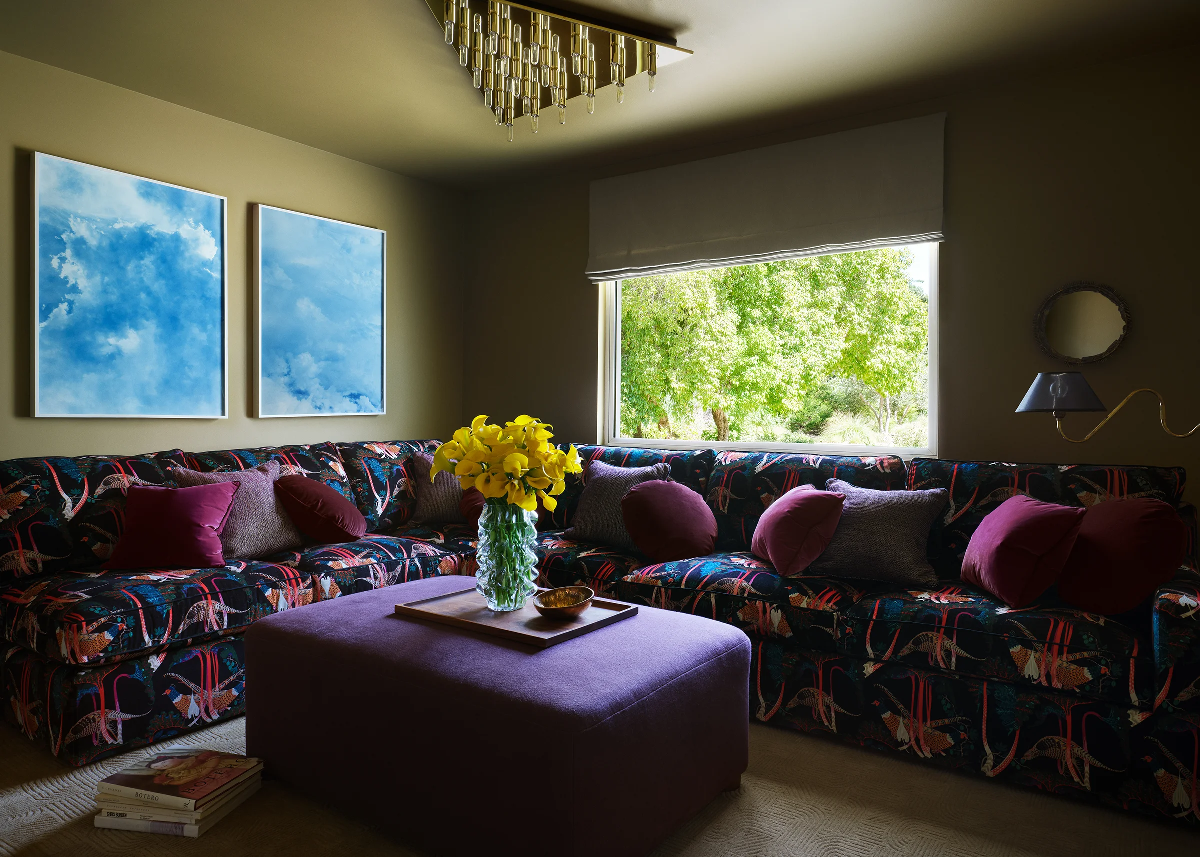 A bold patterned sectional sofa with vibrant botanical upholstery and jewel-toned pillows lounges beneath cloud paintings and brass chandelier with garden views in eclectic Sonoma den.