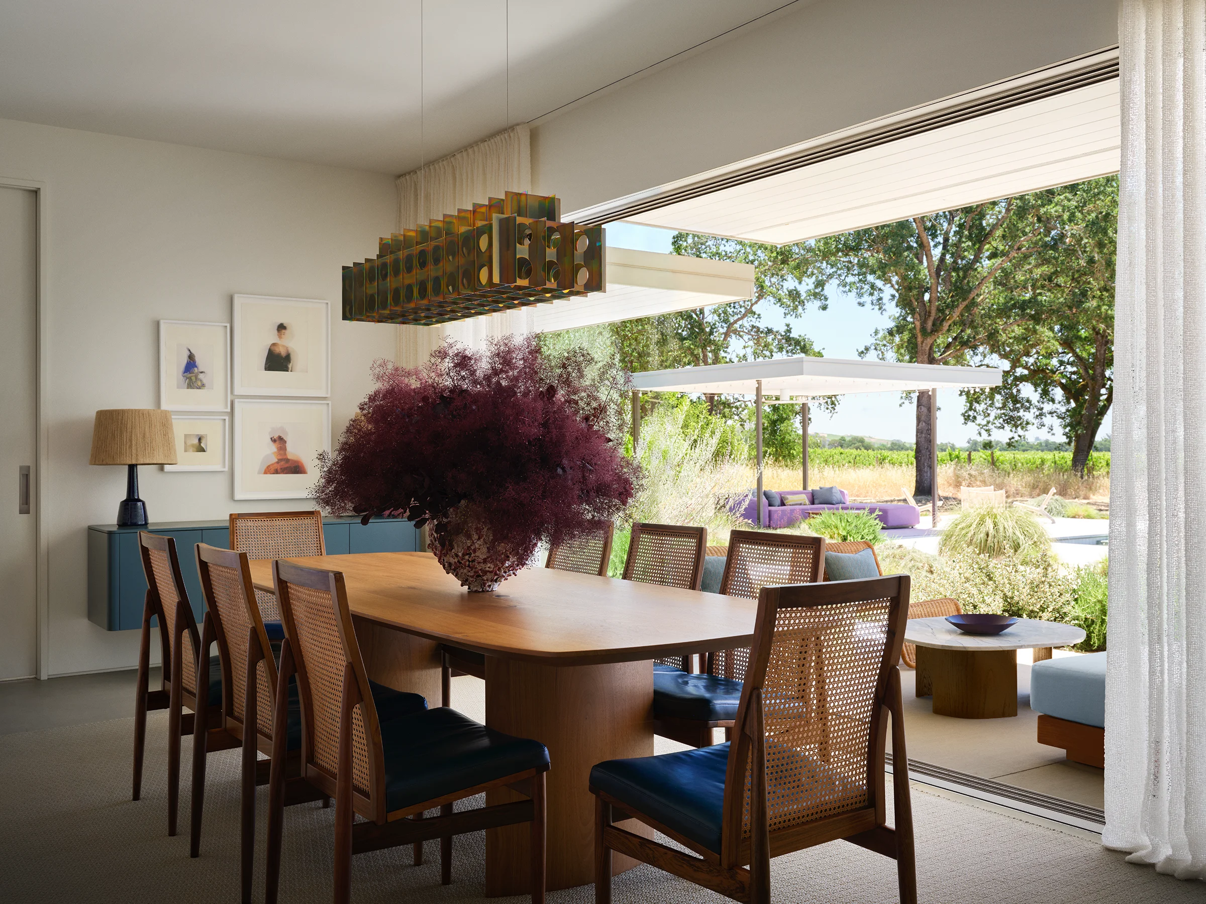 Vintage cane dining chairs with teal leather cushions gather at a wood table beneath a sculptural pendant light with vineyard views and outdoor lounge beyond in Sonoma retreat by Chroma with landscape design by Lucy McFadden.