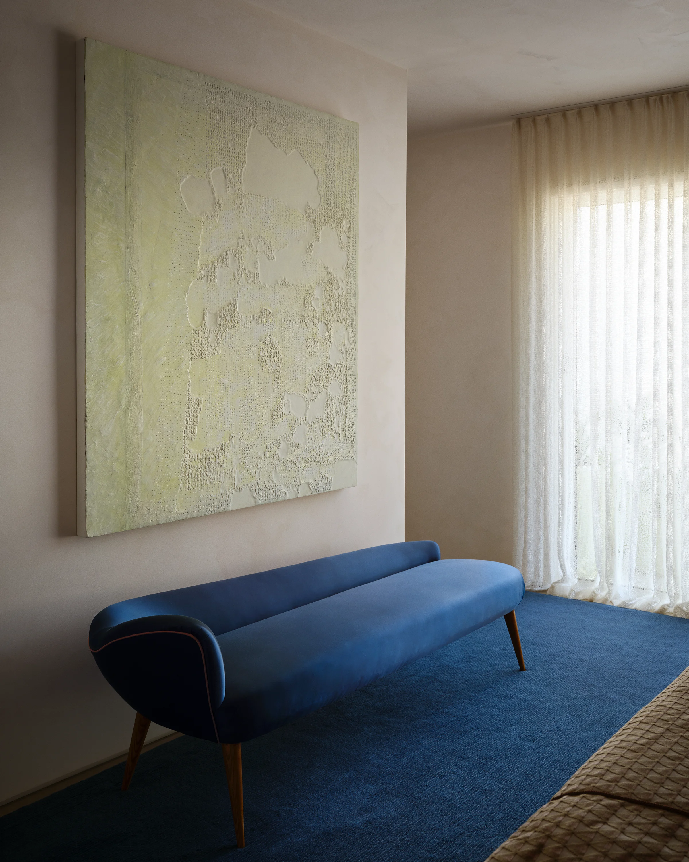 A contemporary abstract artwork in soft yellow and green tones anchors a blue velvet bench on coordinating carpet in minimal Sonoma bedroom.