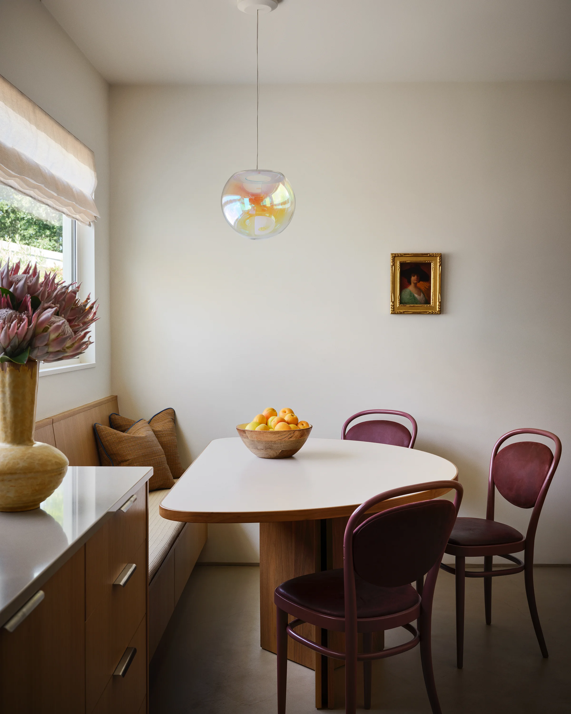 An intimate breakfast nook with burgundy bentwood chairs, iridescent glass pendant, and antique portrait frames garden views in contemporary Northern California vacation home.