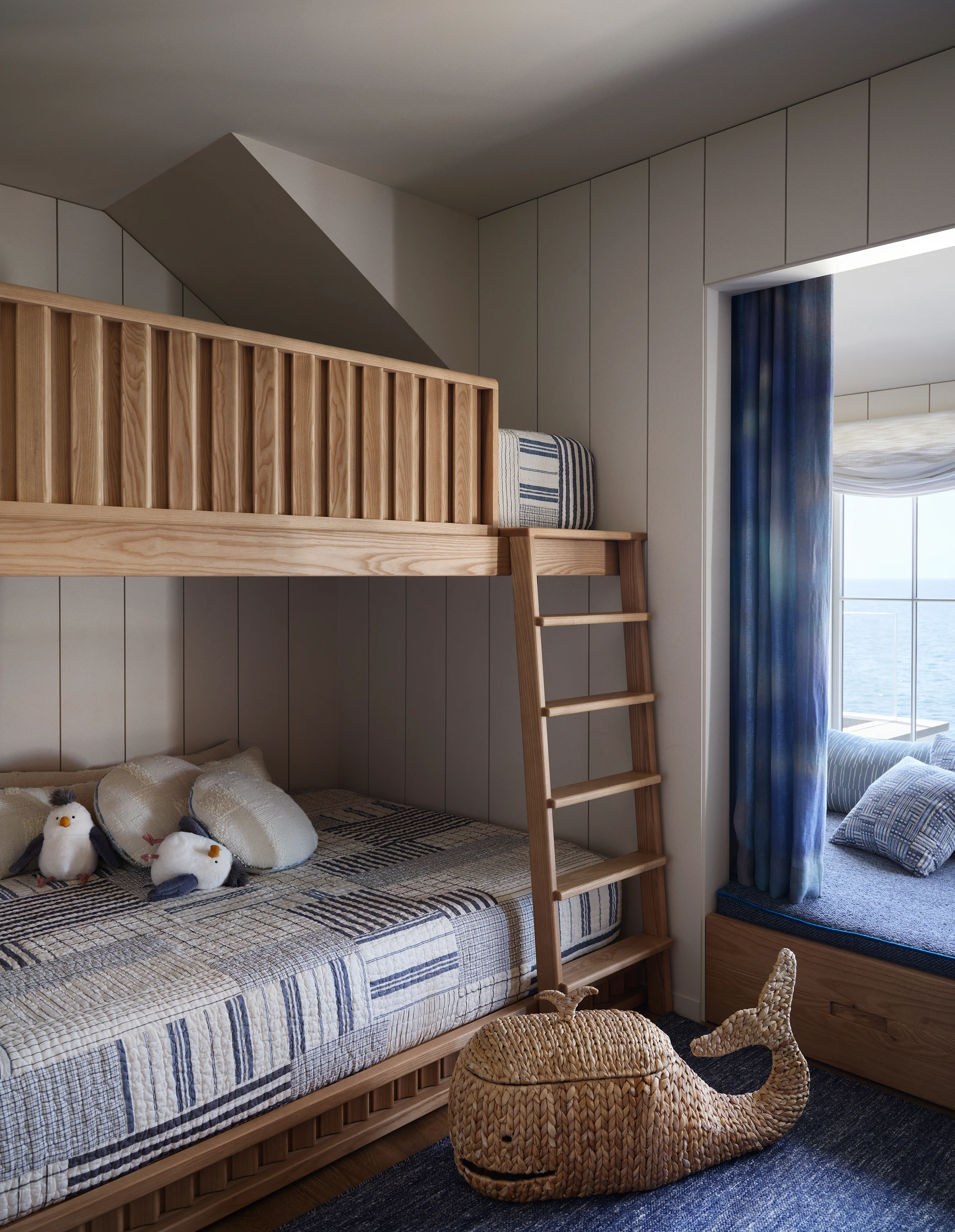 Children's bedroom bunk beds in natural ash with striped quilts, woven whale toy chest, and ocean-view window seat alcove in Santa Cruz home by Chroma.