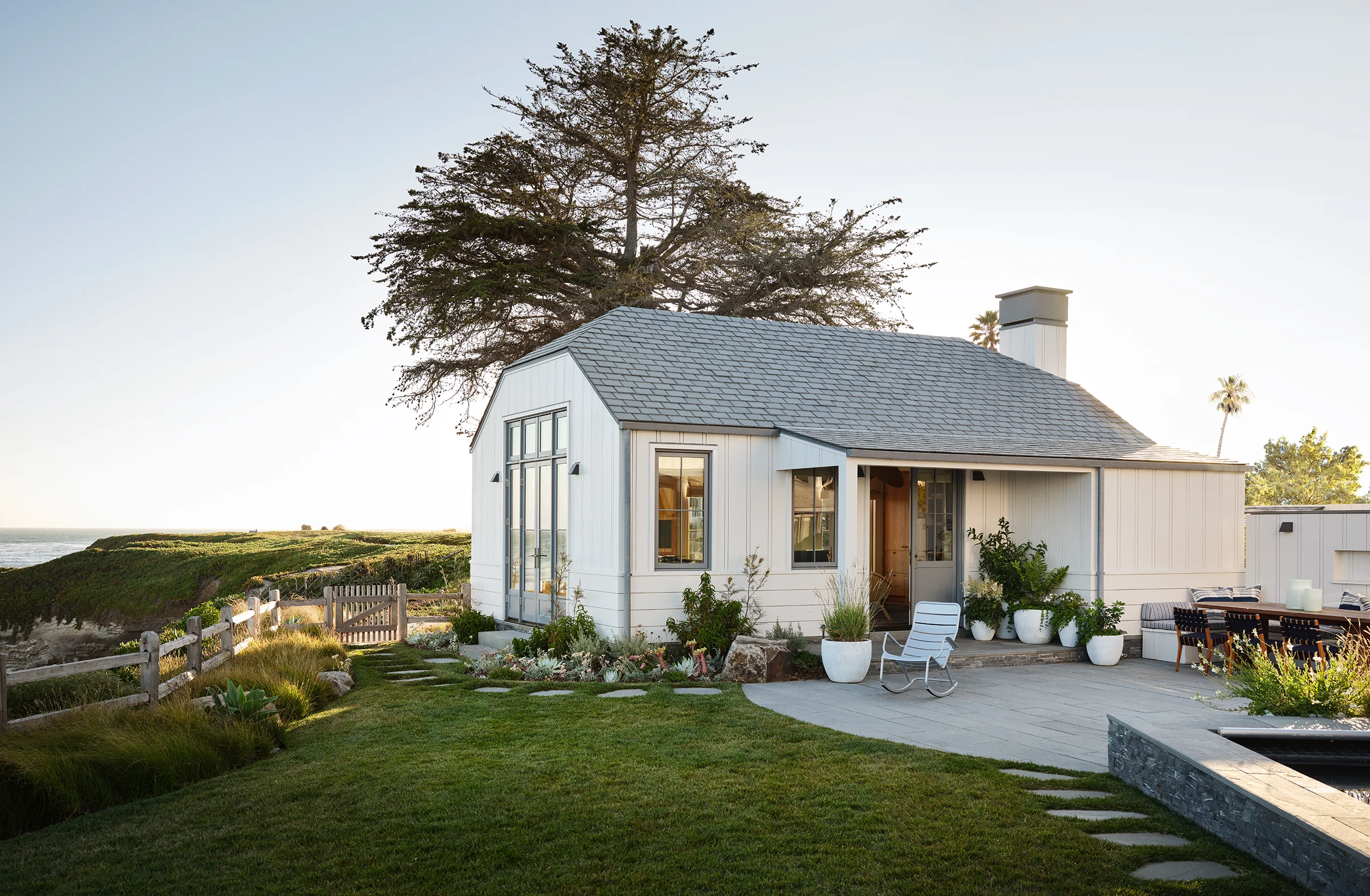 Casita exterior at dusk with board-and-batten siding, Monterey cypress, and garden terrace connecting to main cottage at Santa Cruz blufftop property by Chroma and McGriff Architects.