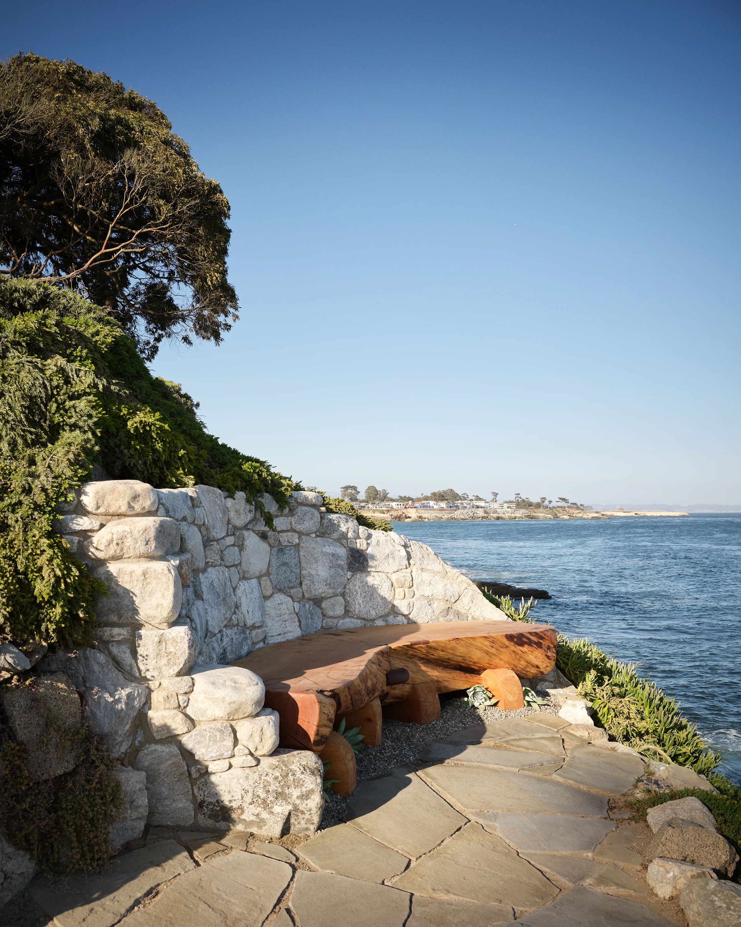 Cliffside stone terrace with hand-hewn cypress log bench against rough granite sea wall overlooking Monterey Bay at the Santa Cruz cottage designed by Chroma.