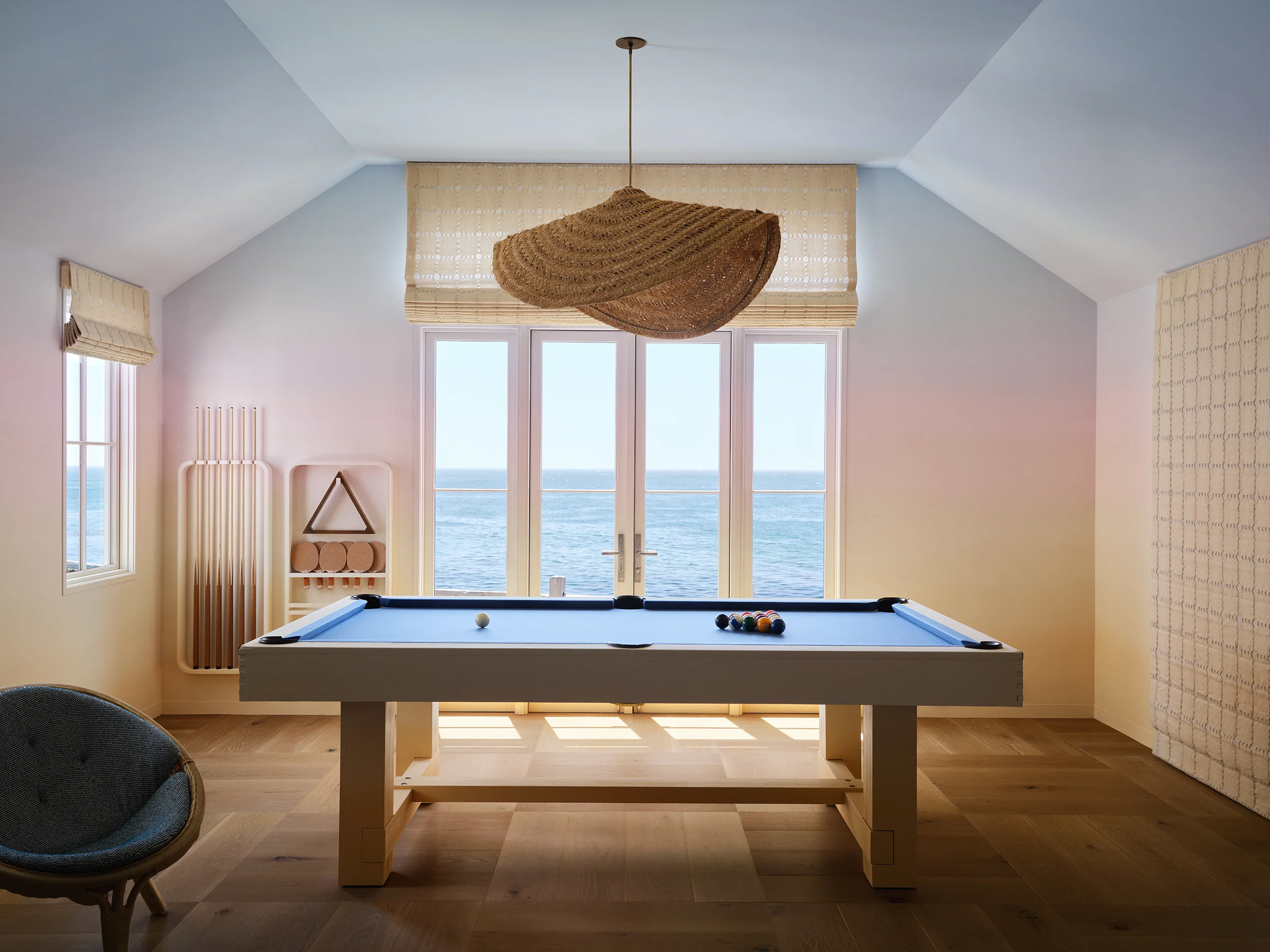 Game room in casita with pool table, woven rattan pendant, blush pink-blue sunset gradient walls, and ocean view French doors in Santa Cruz home by Chroma.