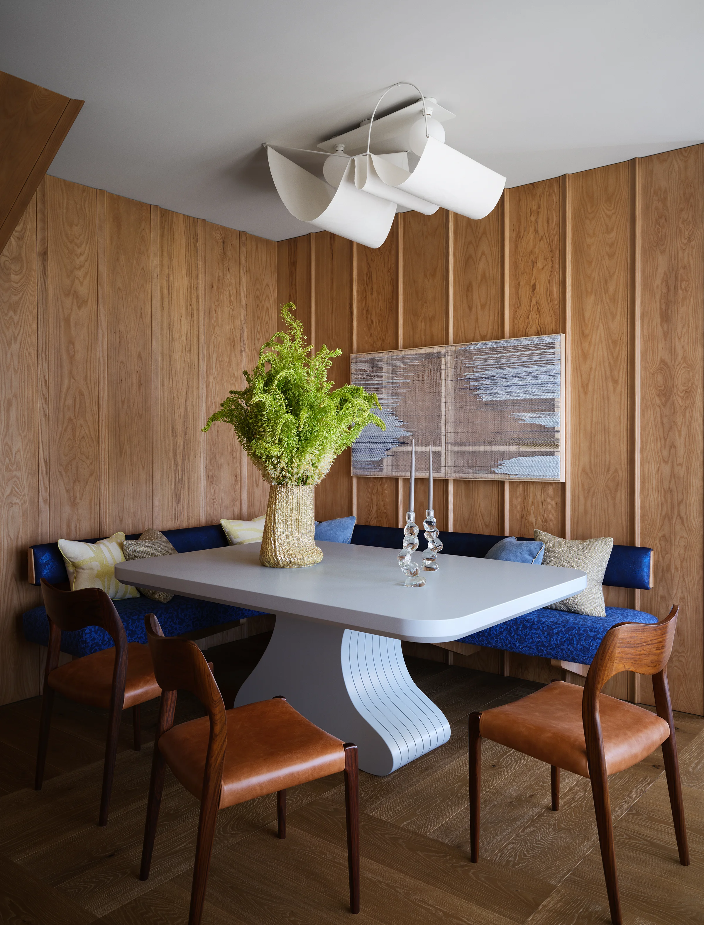 Dining room with wave-inspired wood paneling, wave-form white table on sculptural base, navy built-in banquette, Danish leather chairs, and undulating ceiling fixture in Santa Cruz home by Chroma.