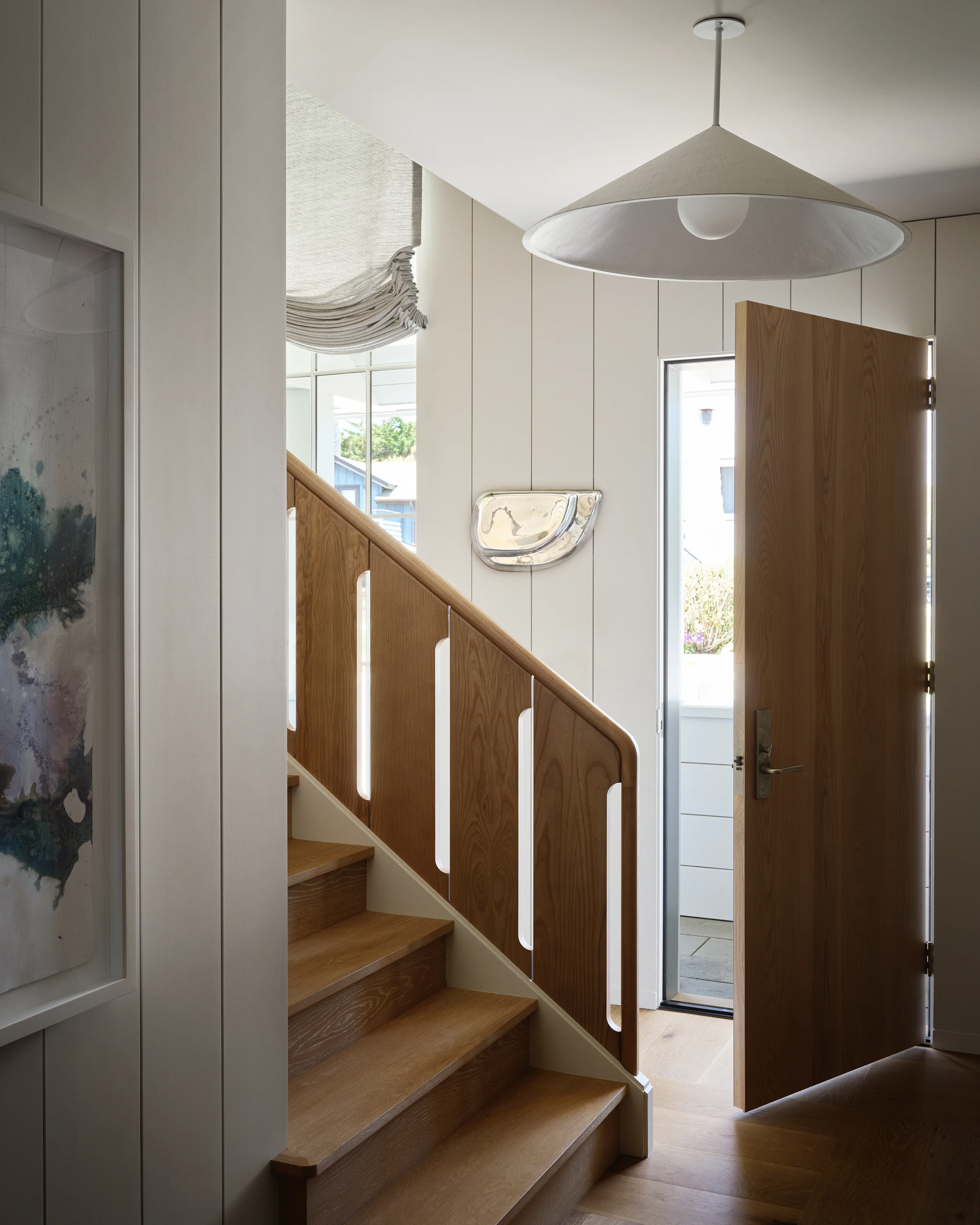 Warm oak staircase with rounded cutout banister panels, plaster pendant light, and metallic ceramic wall sculpture in entry hall of Santa Cruz coastal home by Chroma.