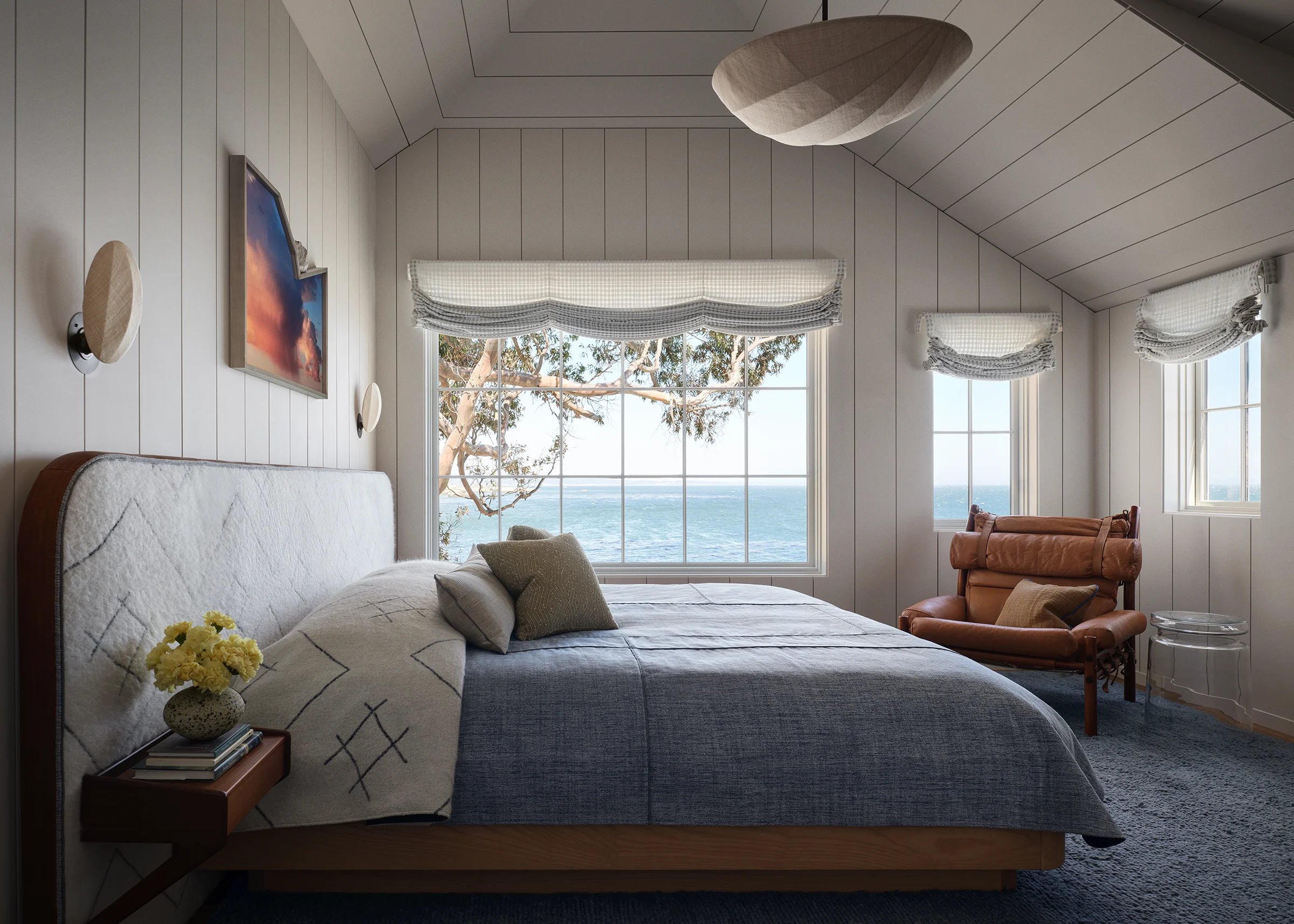 Primary bedroom with slatted white paneled walls, linen pendant, quilted upholstered headboard, blue carpet, ocean panorama through picture windows and open terrace doors in Santa Cruz home by Chroma.