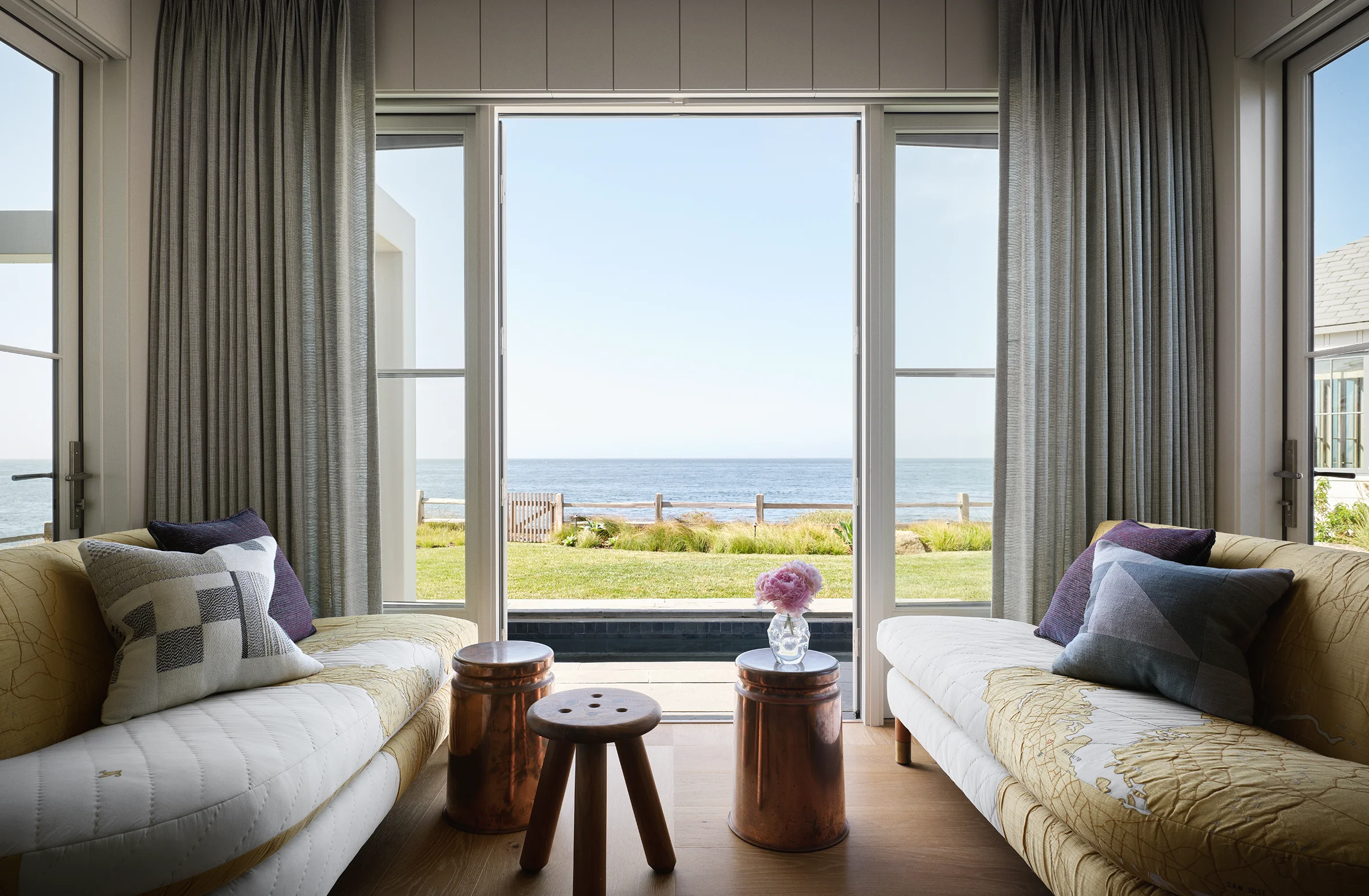 Pair of map-quilted daybeds flanking open ocean doors with copper garden stools in reading room of Santa Cruz blufftop home by Chroma.
