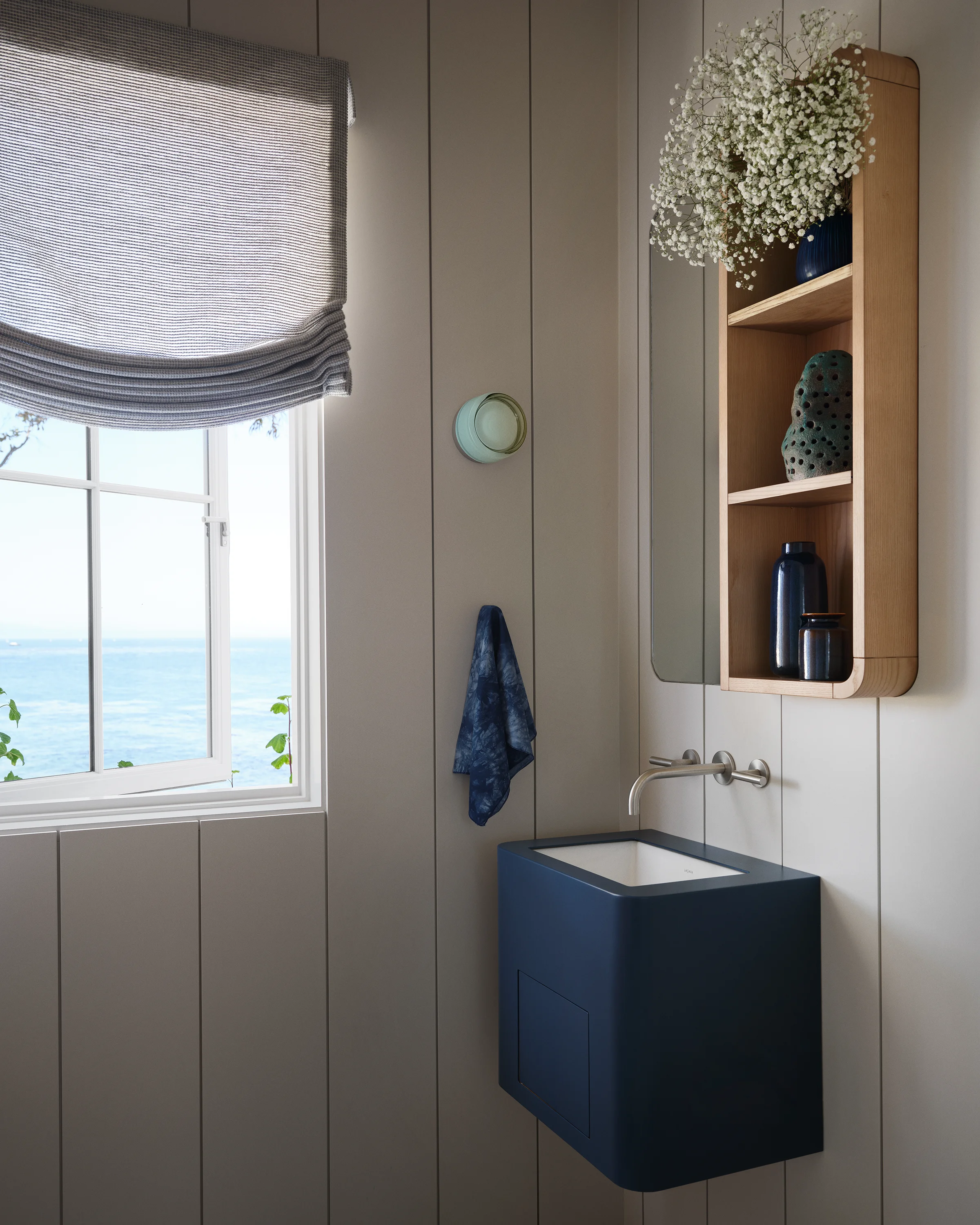 Half bath with navy wall-mounted ceramic sink, oak medicine cabinet, linen roman shade, and ocean view window in Santa Cruz cottage by Chroma.