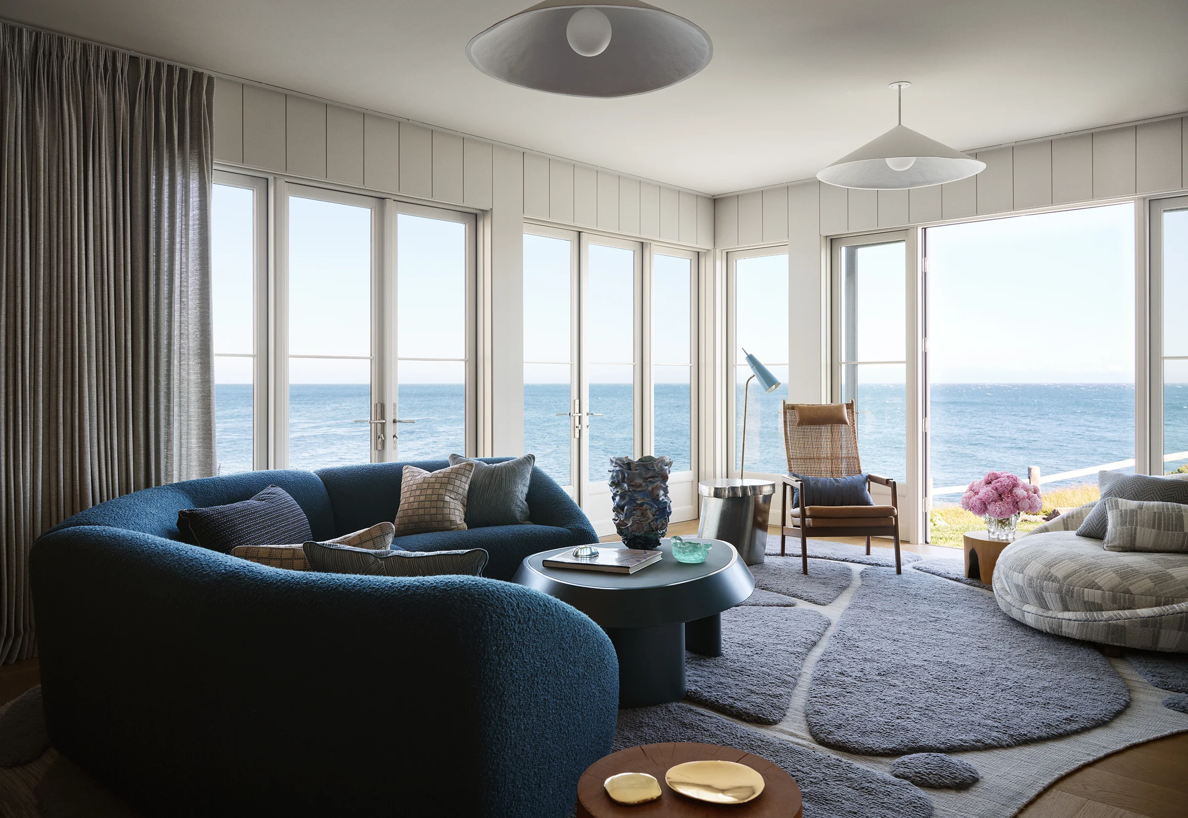 Ocean-facing living room with curved navy bouclé sectional, tide-pool-inspired custom rug, vintage cane chair, and floor-to-ceiling windows overlooking Monterey Bay in Santa Cruz home by Chroma.