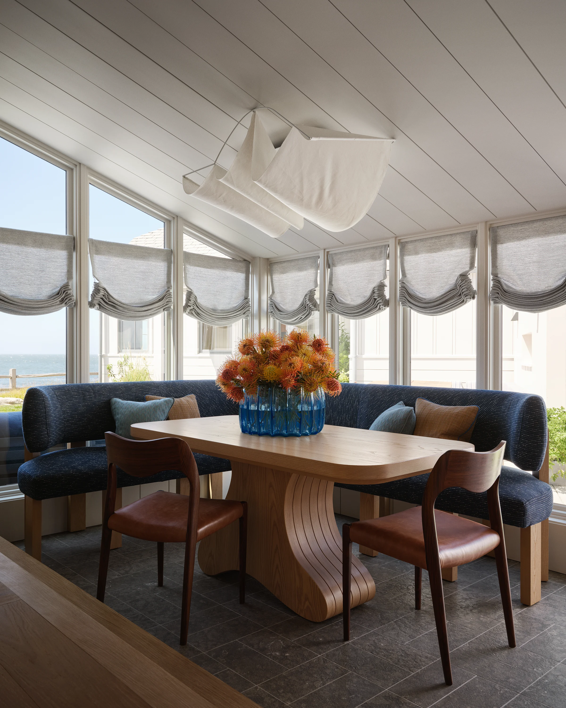Sun-drenched breakfast nook with curved navy banquette, surfboard-shaped wood table, sculptural fabric ceiling pendant, and roman shades framing panoramic Monterey Bay views in Santa Cruz home by Chroma.