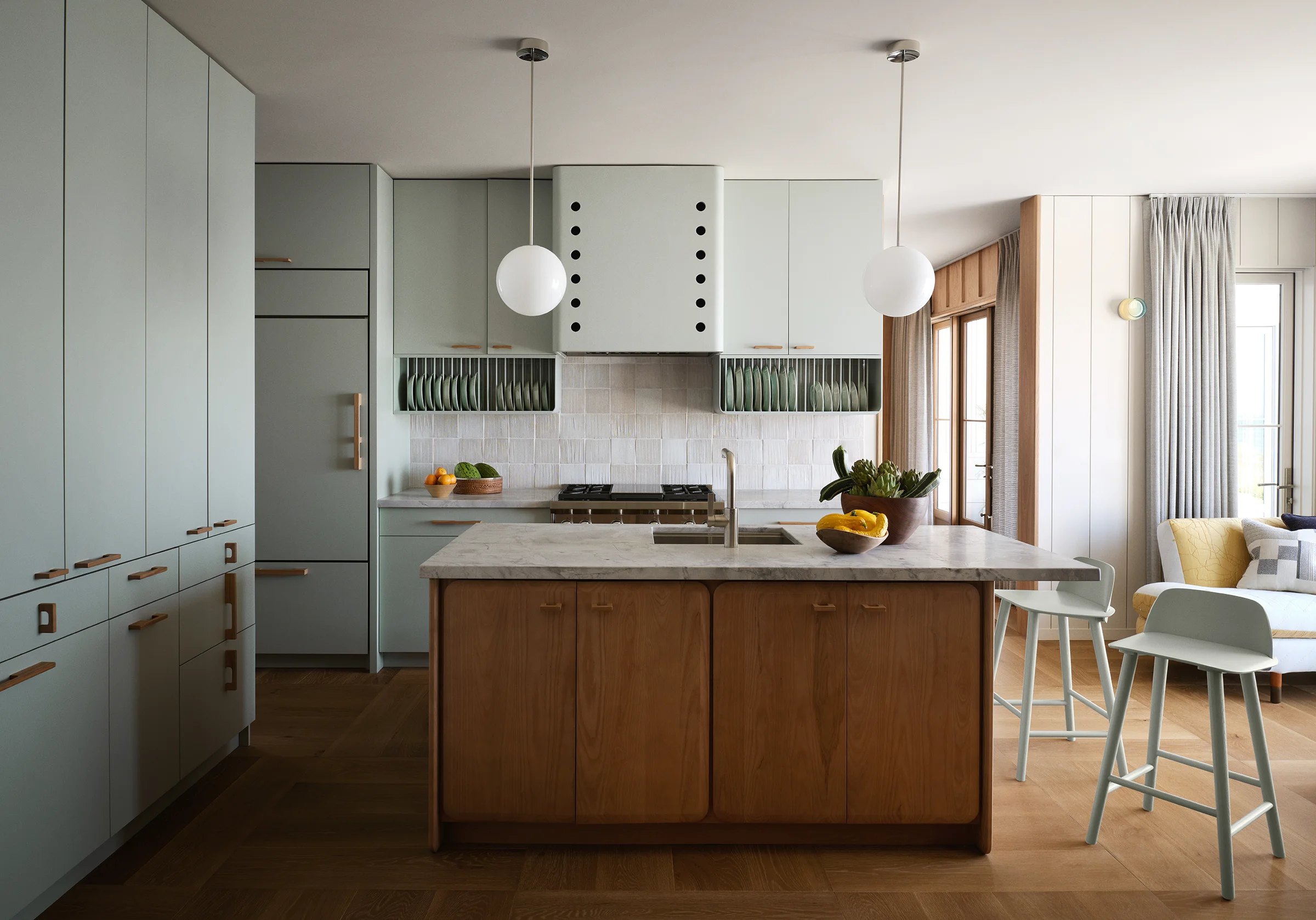 Sage green kitchen with custom rounded hood, globe pendants, open plate racks, wood island with marble countertop, and herringbone floors in Santa Cruz coastal home by Chroma.