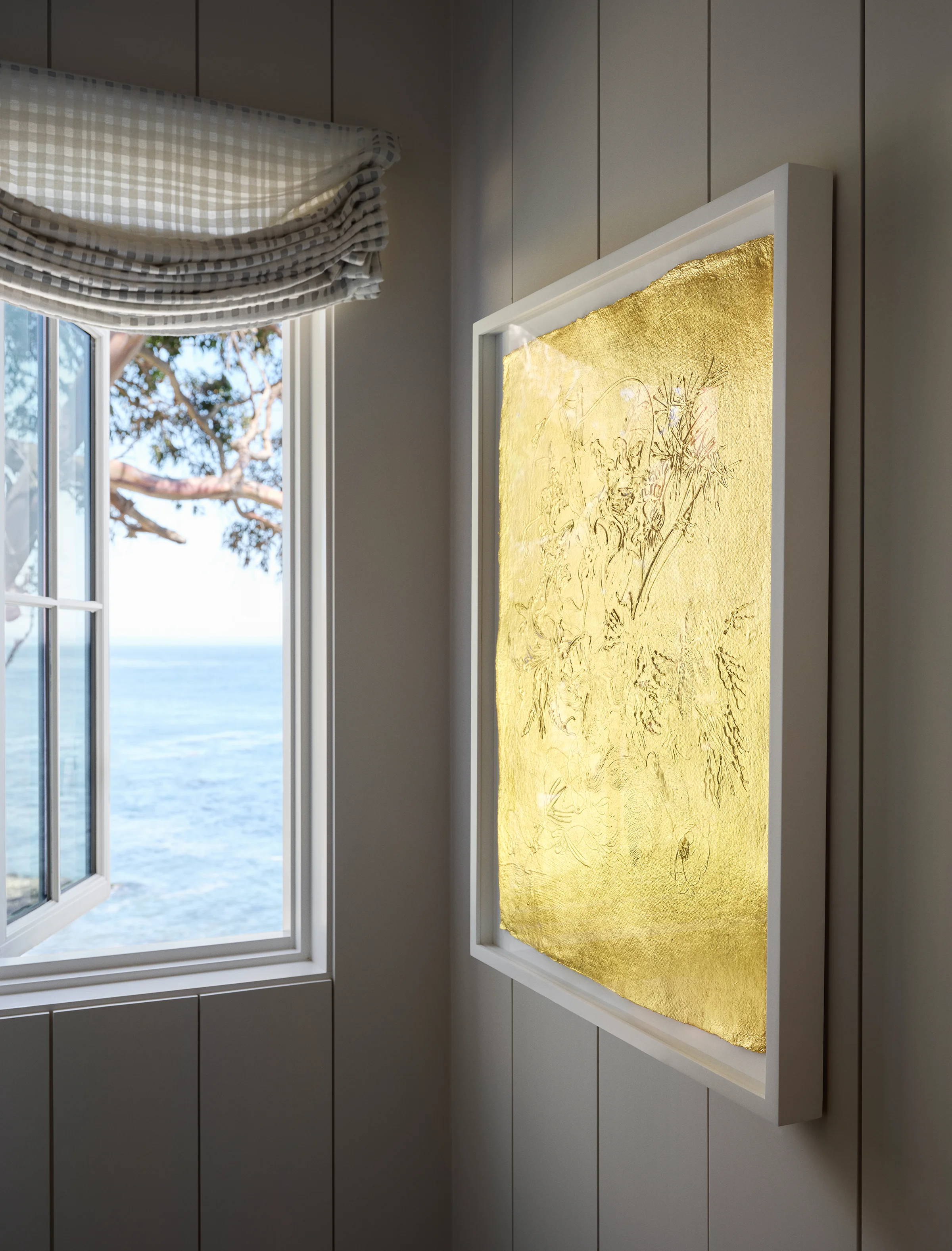 Illuminated gold-leaf botanical artwork glowing beside ocean-view window with eucalyptus branches in primary suite of Santa Cruz home by Chroma.
